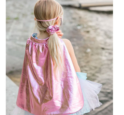 niña de espaldas con la capa de La capa rosa metalizado, forrada con un precioso satén azul, ondea con la elegancia de un superhéroe, haciendo que cada pequeño héroe destaque. El antifaz rosa metalizado a juego añade un aire de misterio, perfecto para ocultar su identidad secreta.