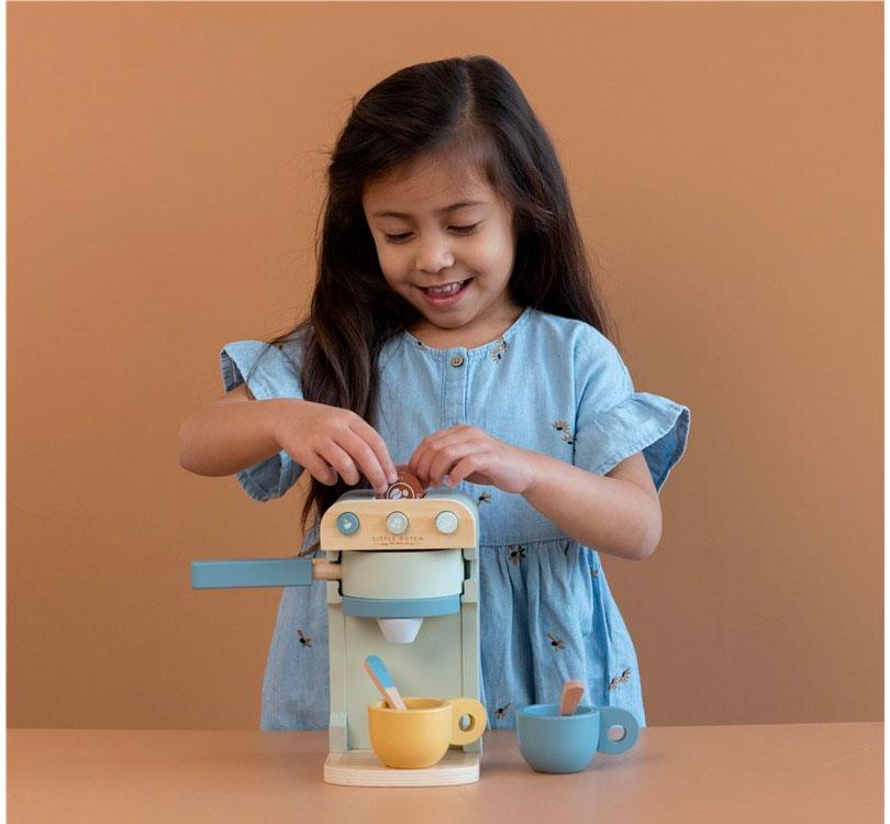 Niña morena de pelo largo jugando con foto de producto de cafetera de madera de la marca Little Dutch en tonos menta, madera natural y combinación con mostaza tanto las tazas como las cápsulas