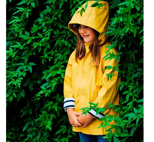 chubasquero Euri en color amarillo brillante diseño  en color amarillo impermeable. Los bolsillos frontales son súper prácticos, y el forro interior a rayas marinero añade un toque de estilo de lo más molón. Foto de niña escondida en un seto con el chubasquero puesto