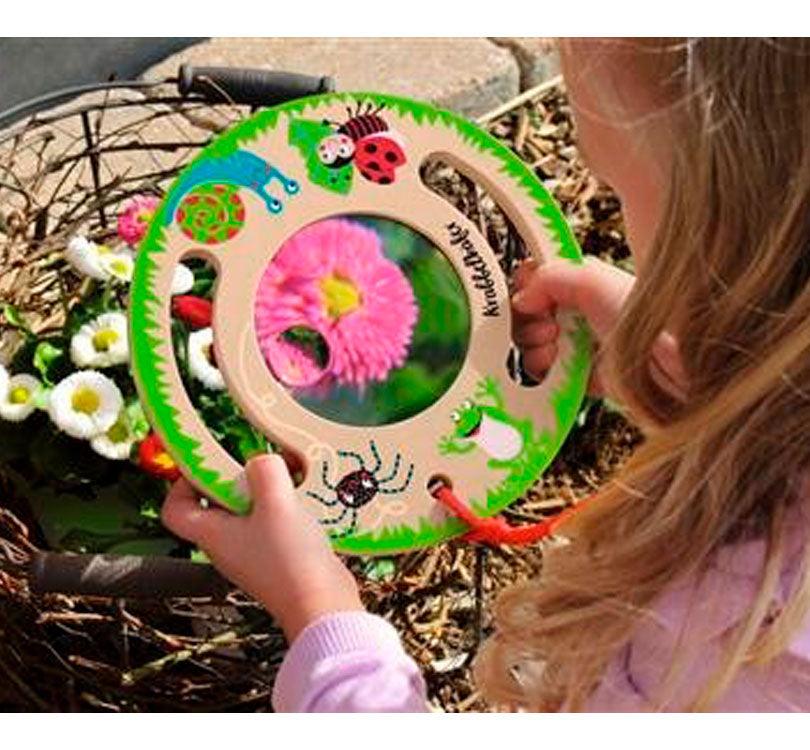 lupa gigante de forma circular de madera con asas para poder ser agarrada de forma ligera por una niña que observa flores