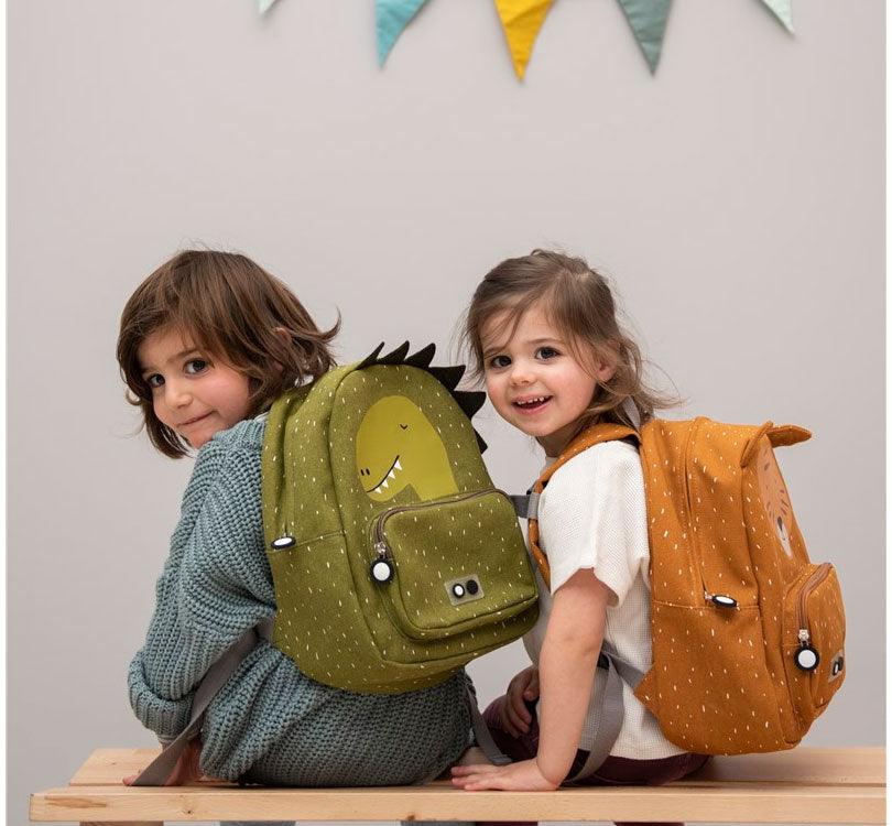 niños con mochilas tirie puestas, una de ellas la Mochila Dino de Trixie Baby ha llegado para convertir la vuelta al cole en una aventura inolvidable! ¡Ideal para la  etapa de infantil!  Esta mochila con forma de Dino, con su carita impresa y sus divertidas escamas, conquistará el corazón de vuestros peques. Sus asas acolchadas y ajustables brindan comodidad en cada paso, mientras el cierre con clip en el pecho evita cualquier percance. color verde kakhi