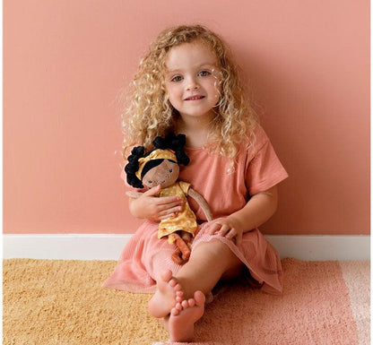 niña rubia de pelo rizado y largo sentada en el suelo jugando con muñeca blandita evi de Little Dutch de piel morena con cabello rizado negro vestido estampado mostaza y botas color teja 