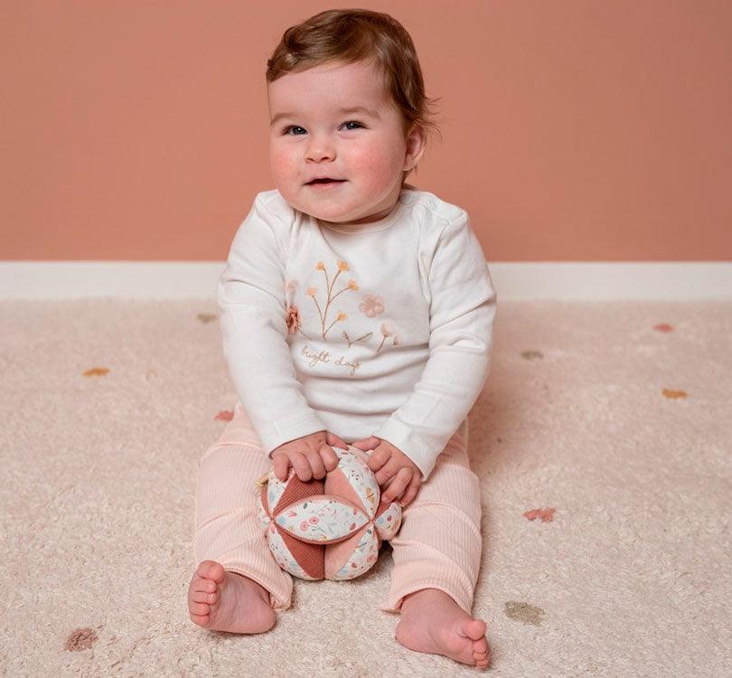 niña sentada en el suelo en una habitación de tonos rosas con la bola de agarra sensorial o pelota montessori de la colección flores y mariposas de la marca Little Dutch en colores rosas y con estampados de flores 