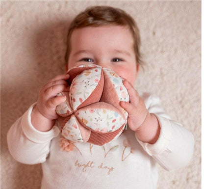 Bebé en primer plano con la niña sentada en el suelo en una habitación de tonos rosas con la bola de agarra sensorial o pelota montessori de la colección flores y mariposas de la marca Little Dutch en colores rosas y con estampados de flores 