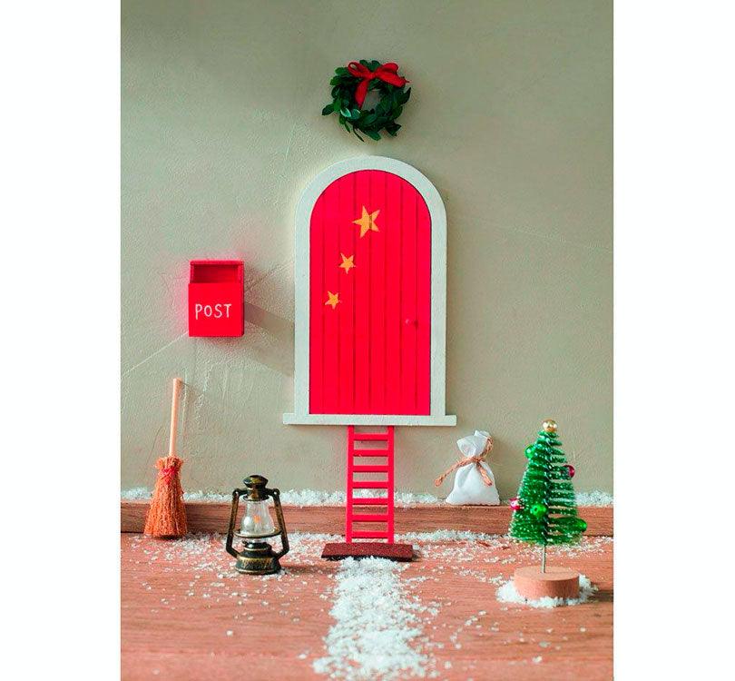 foto lifestyle de la adorable puerta decorativa! Hecha de madera y plástico, esta pequeña puerta  incluye 9 encantadoras piezas navideñas, ¡como un buzón, una escalerita, una escoba y más! color rojo marca Moses 