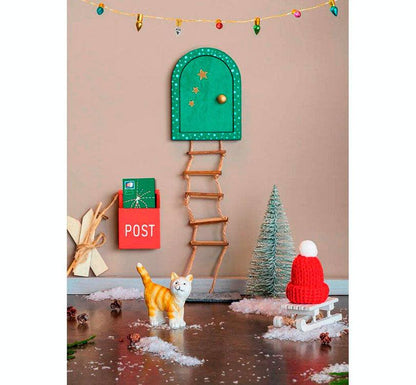 foto de lifestyle de la adorable puerta decorativa! Hecha de madera y plástico, esta pequeña puerta  incluye 10 encantadoras piezas navideñas, ¡como un buzón, un gatito, un trineo y más! color verde marca Moses