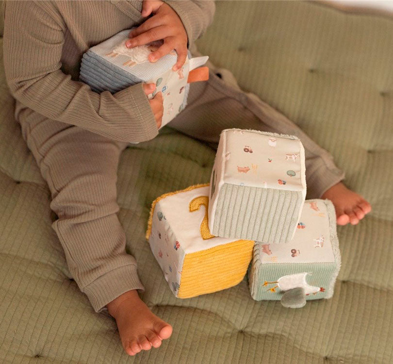 niña jugando onjunto de cubos blanditos de la colección Pequeña Granja.  Estimula la coordinación mano-ojo de tu pequeño mientras se divierte durante horas. Con 4 cubos sensoriales, cada uno diseñado con colores suaves y un número único, tu bebé también podrá explorar divertidas etiquetas, caras de felpa y un espejo soleado.