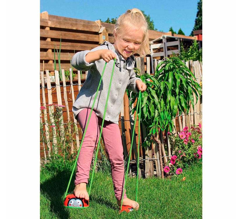 niña rubia en el jardín usando zancos de madera con forma circular y estampado de mariquitas, con cuerdas ajustables en verde lima de la marca Moses