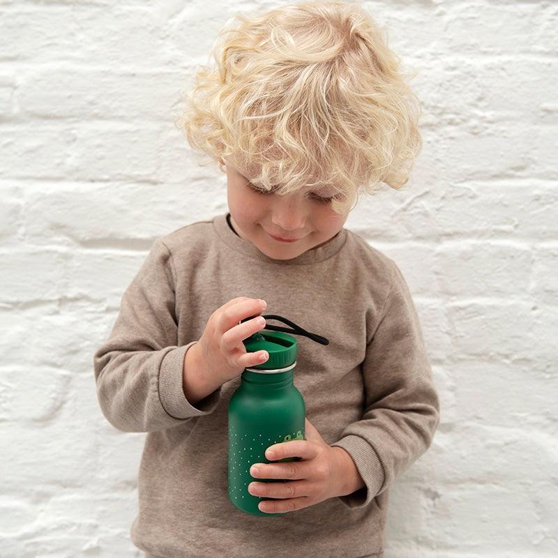 foto de lifestyle de un niño muy rubio apoyado en pared blanca con la BOTELLA DE ACERO DE LA MARCA TRIXIE DE FORMA CILINDRICA CON TAPON DE PITORRO AL TONO, MODELO COCODRILO EN COLOR VERDE