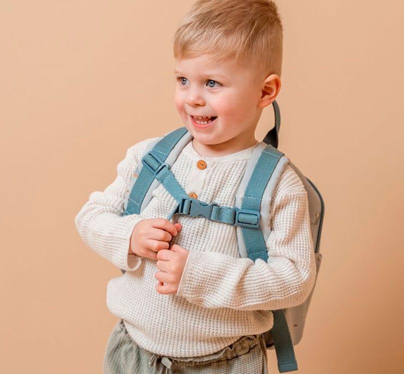 niño rubio con la mochila pequeña para etapa de preescolar de la marca Little Dutch con estampado marinero en tono azul claro  y cremalleras en tono azul grisáceo con un bolsillo grande de cremallera y uno pequeño frontal también de cremallera abrochada al pecho con las tras en tono azulon