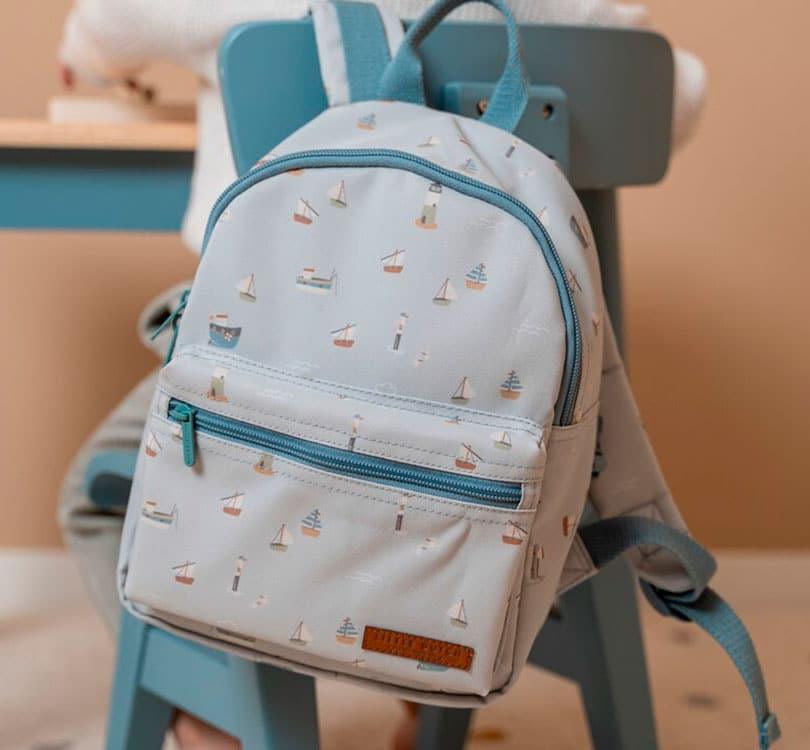 colgada de un asa sobre una silla azul vemos la mochila pequeña para etapa de preescolar de la marca Little Dutch con estampado marinero en tono azul claro  y cremalleras en tono azul grisáceo con un bolsillo grande de cremallera y uno pequeño frontal también de cremallera